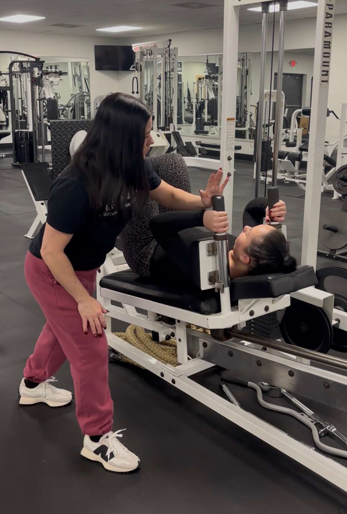 Female Personal Trainer assisting female client with a leg press machine in a gym.